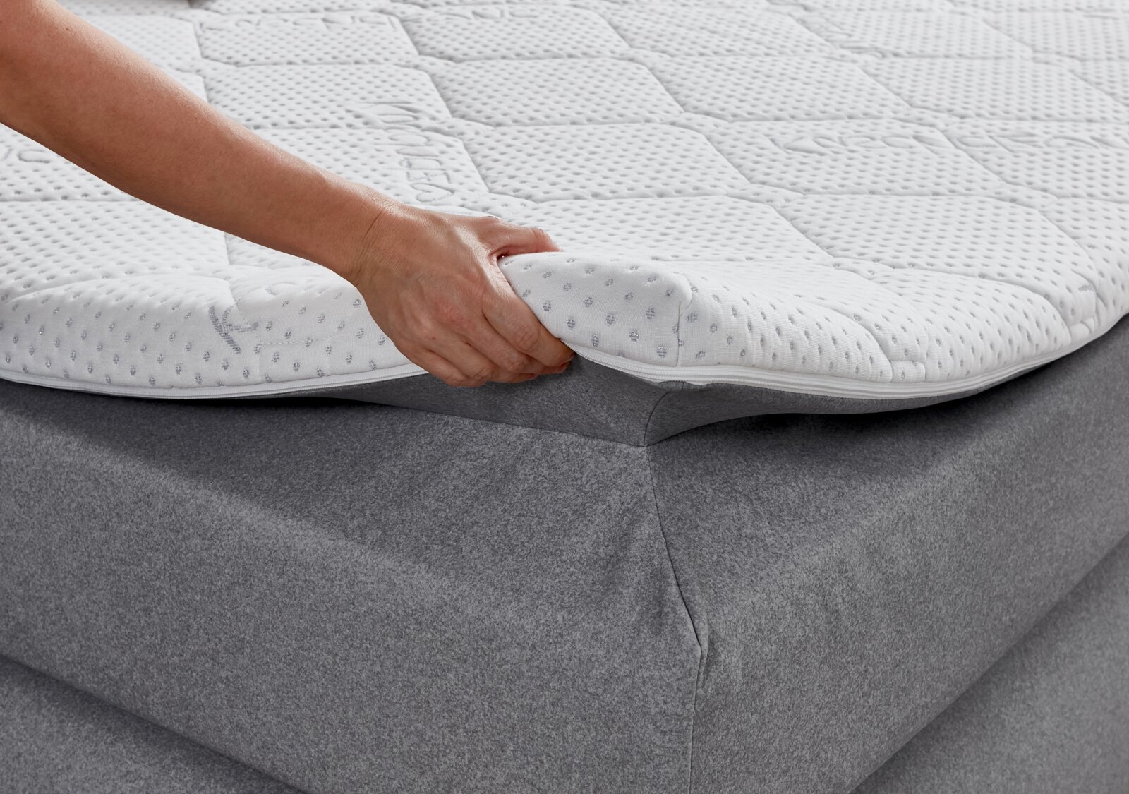 Nahaufnahme eines Boxspringbetts mit einer Hand, die den Matratzentopper anhebt, um die weiche, gepolsterte Oberfläche zu zeigen.