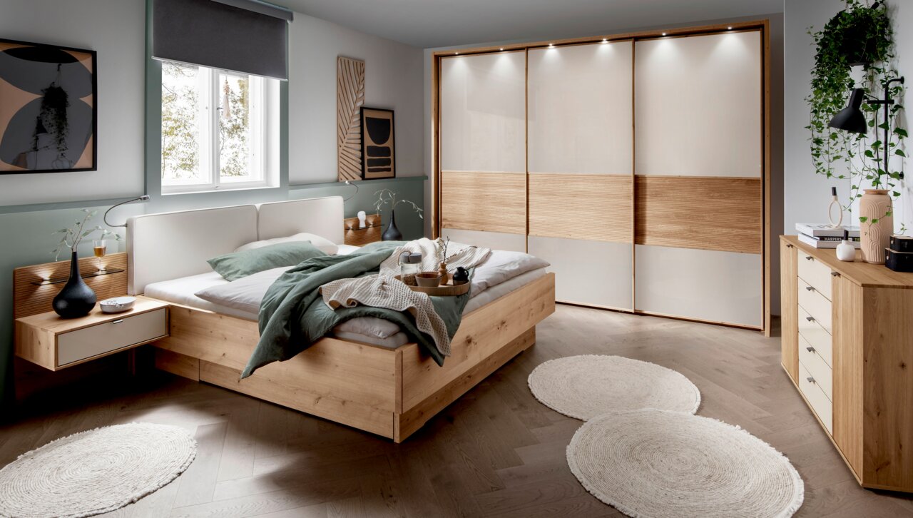 Modernes Schlafzimmer mit einem Futonbett aus Holz, seitliche Perspektive. Das Bett ist mit grüner Bettwäsche und Kissen dekoriert, umgeben von stilvollen Nachttischen und einem großen Kleiderschrank mit Schiebetüren.