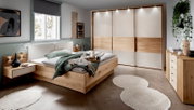 Modernes Schlafzimmer mit einem Futonbett aus Holz, seitliche Perspektive. Das Bett ist mit grüner Bettwäsche und Kissen dekoriert, umgeben von stilvollen Nachttischen und einem großen Kleiderschrank mit Schiebetüren.