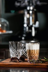 Zwei elegante Barista Kaffeebecher aus Glas, einer gefüllt mit Cappuccino, auf einem Holzbrett mit Schokoladenstücken und Kräutern, seitliche Perspektive.