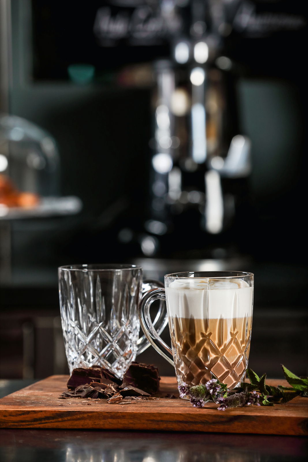 Zwei elegante Barista Kaffeebecher aus Glas, einer gefüllt mit Cappuccino, auf einem Holzbrett mit Schokoladenstücken und Kräutern, seitliche Perspektive.