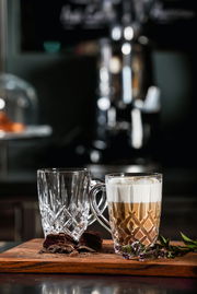 Zwei elegante Barista Kaffeebecher aus Glas, einer gefüllt mit Cappuccino, auf einem Holzbrett mit Schokoladenstücken und Kräutern, seitliche Perspektive.