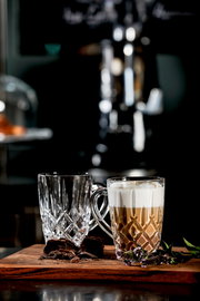 Zwei elegante Barista Kaffeebecher aus Glas, einer gefüllt mit Cappuccino, auf einem Holzbrett mit Schokoladenstücken und Kräutern, seitliche Perspektive.