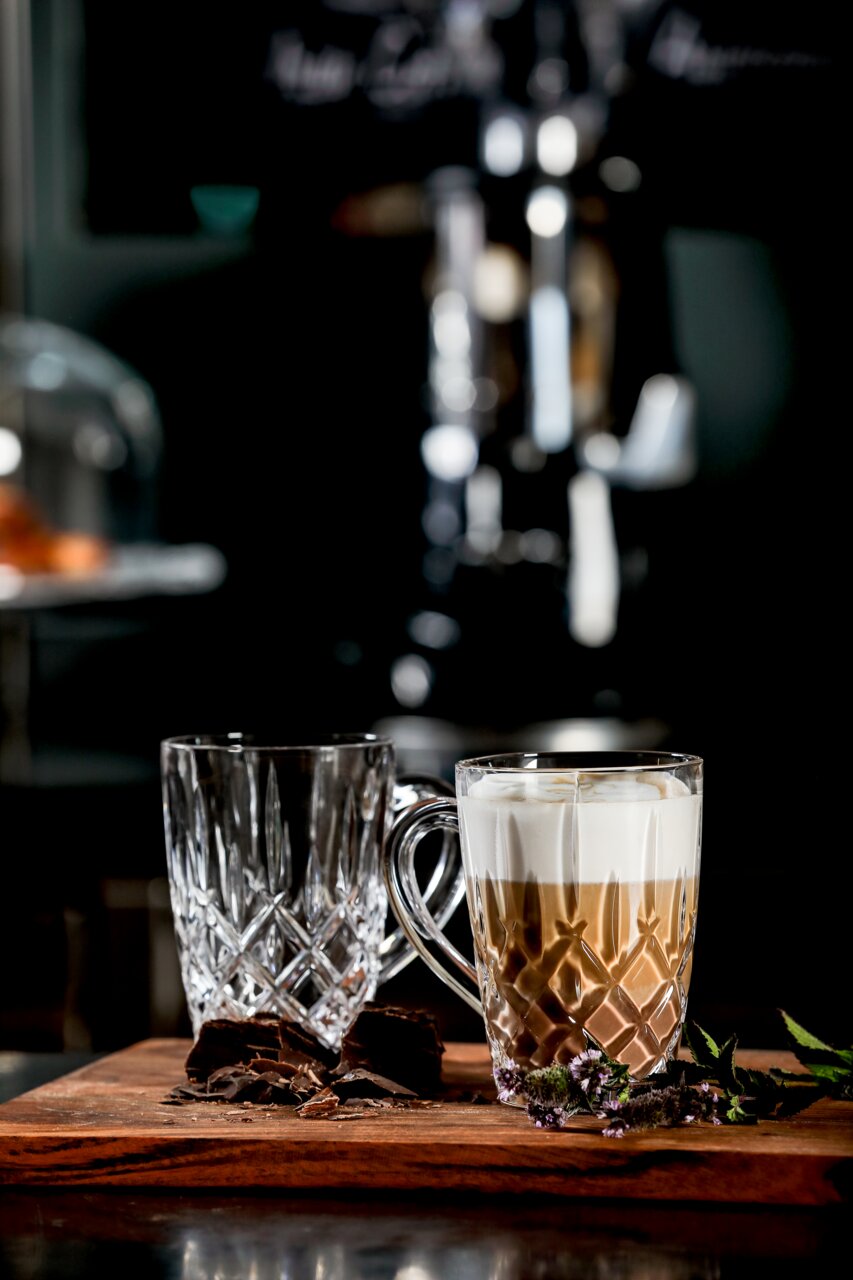 Zwei elegante Barista Kaffeebecher aus Glas, einer gefüllt mit Cappuccino, auf einem Holzbrett mit Schokoladenstücken und Kräutern, seitliche Perspektive.