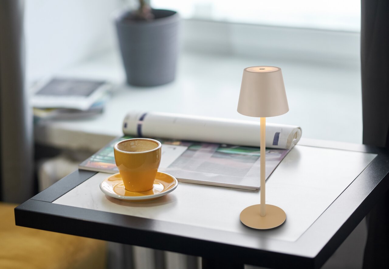 De VANNIE 3-tafellamp op accu in beige op een tafel naast een kopje koffie, gefotografeerd vanuit een zijdelingse hoek.