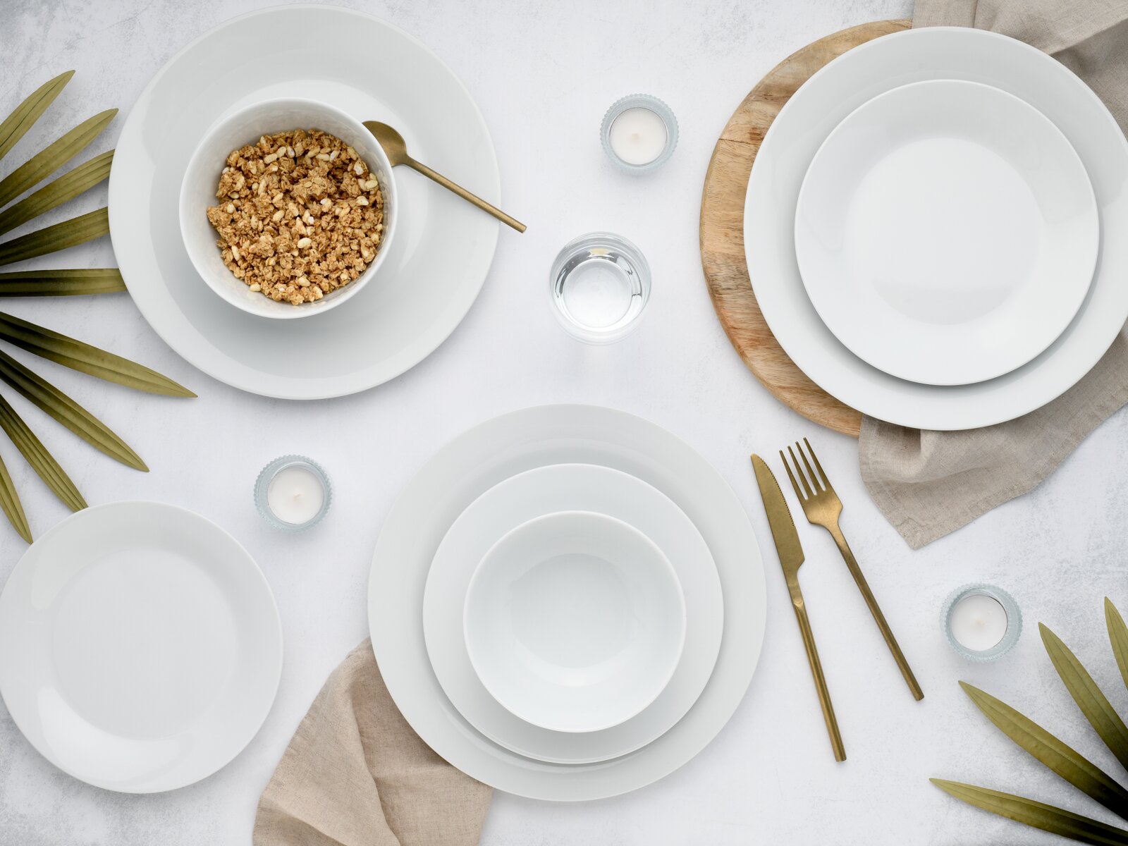Minimalistisches weißes Porzellan-Geschirrset mit Tellern und Schalen, goldfarbenem Besteck, Kerzen und Leinenservietten auf hellem Tisch – Draufsicht von oben.