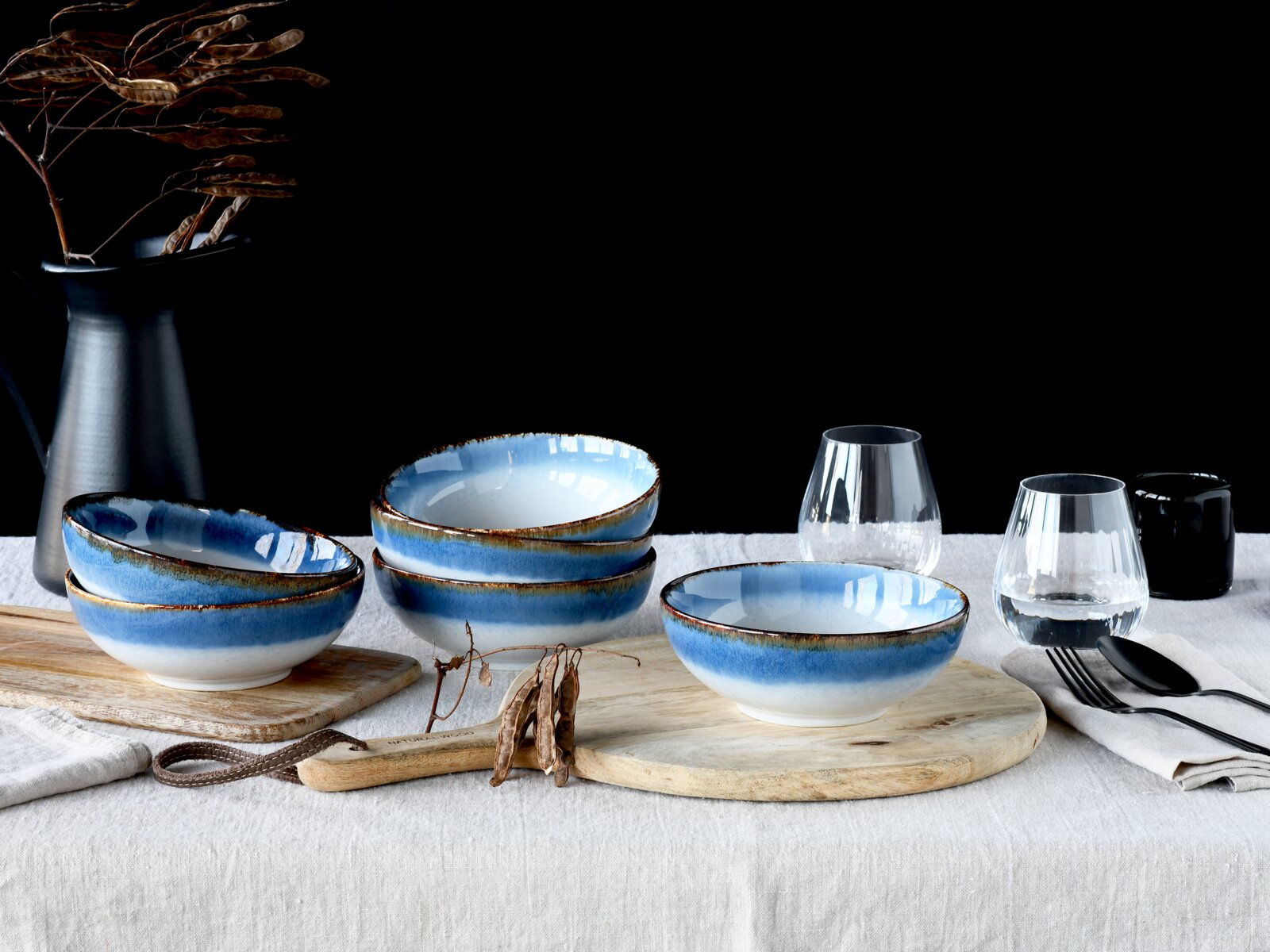 Blau-weiß glasierte Keramikschalen mit braunem Rand auf gedecktem Tisch mit Holzbrettchen, Wassergläsern und Besteck vor schwarzem Hintergrund, perspektivisch in leichter Seitenansicht von vorne