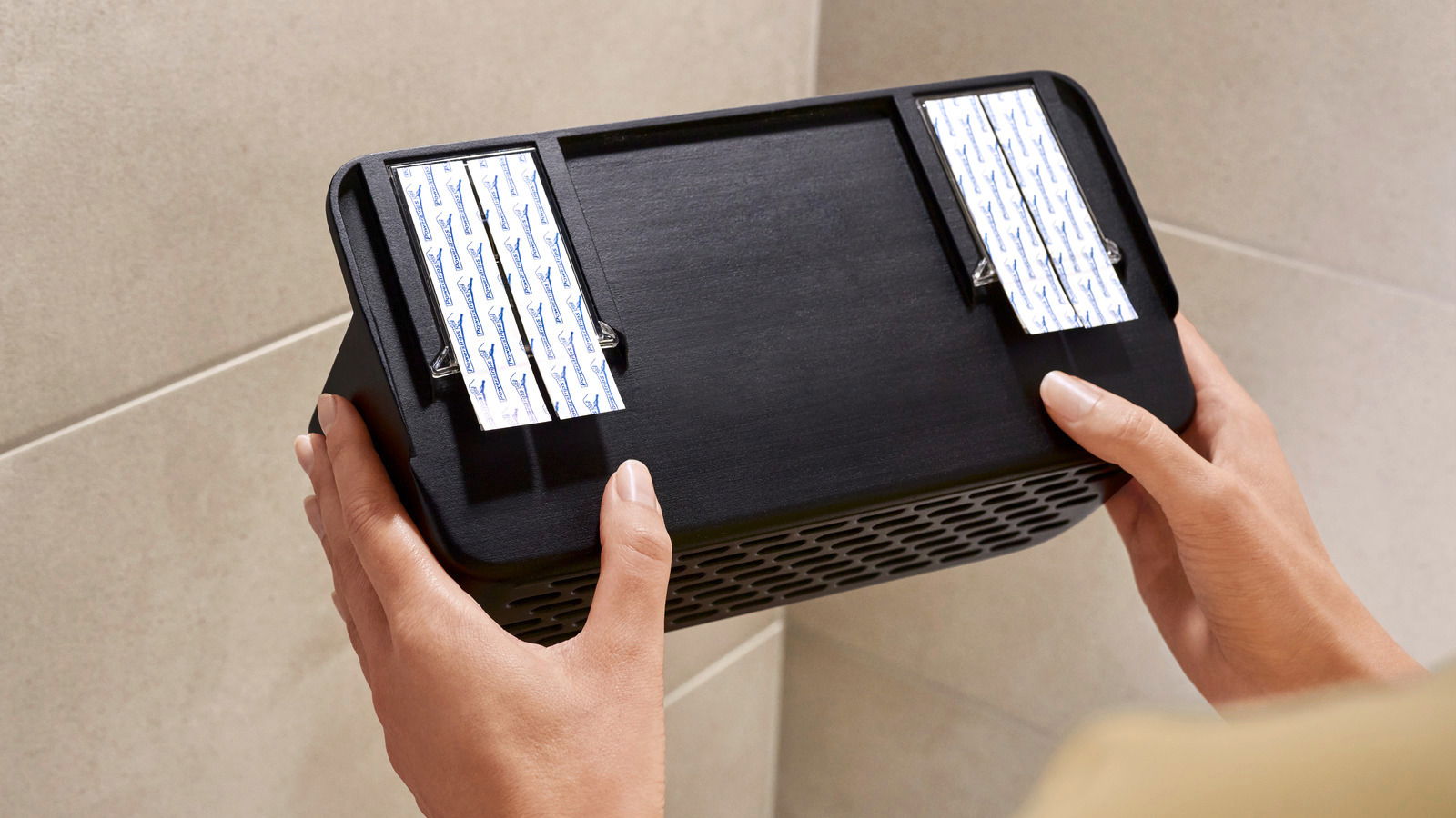 Nahaufnahme eines schwarzen Duschkorbs von tesa Lavaa S, der von zwei Händen gehalten wird. Der Korb wird an einer Wand mit Klebestreifen befestigt.