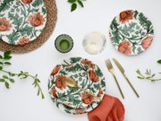 Draufsicht (Flatlay) auf gedeckten Tisch mit weißem Keramikgeschirr mit orangefarbenem Blumen- und grünem Blättermuster, goldenem Besteck, grünem Glas, Wasserglas, roter Serviette und geflochtenem Platzset auf weißem Hintergrund.