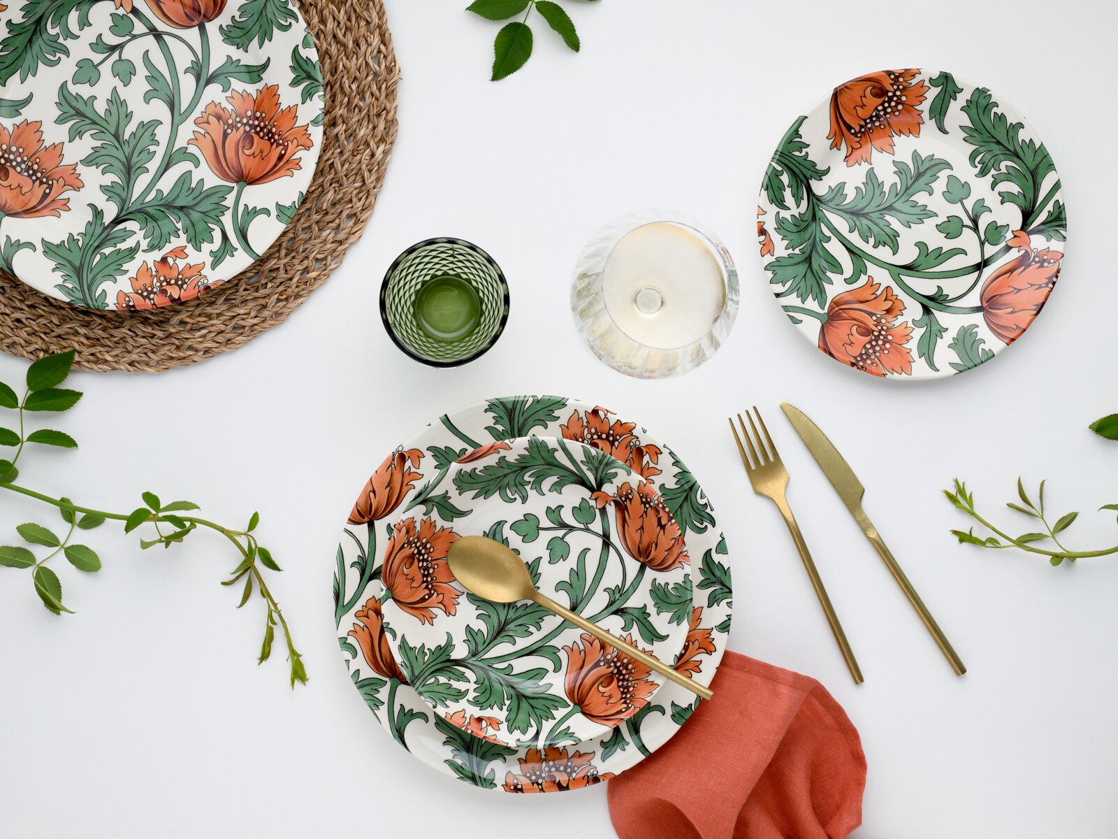 Draufsicht (Flatlay) auf gedeckten Tisch mit weißem Keramikgeschirr mit orangefarbenem Blumen- und grünem Blättermuster, goldenem Besteck, grünem Glas, Wasserglas, roter Serviette und geflochtenem Platzset auf weißem Hintergrund.