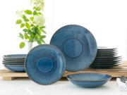 Blau glasiertes Keramik-Geschirrset mit Teller- und Schalenstapeln, ein großer und ein kleiner Teller frontal aufgestellt, aufgenommen in leichter Schrägansicht von vorn auf hellem Tisch vor weißem Hintergrund.