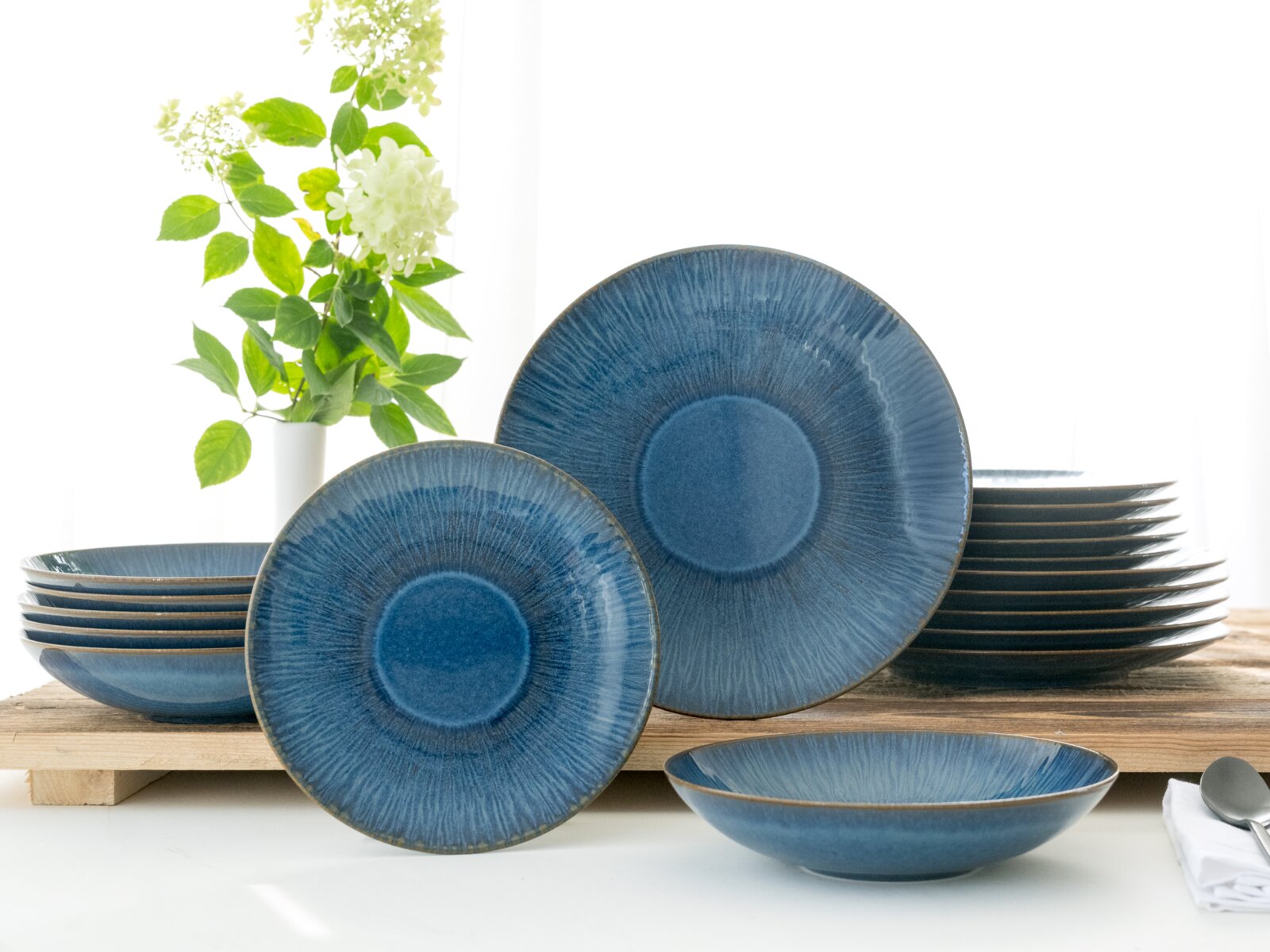 Blau glasiertes Keramik-Geschirrset mit Teller- und Schalenstapeln, ein großer und ein kleiner Teller frontal aufgestellt, aufgenommen in leichter Schrägansicht von vorn auf hellem Tisch vor weißem Hintergrund.