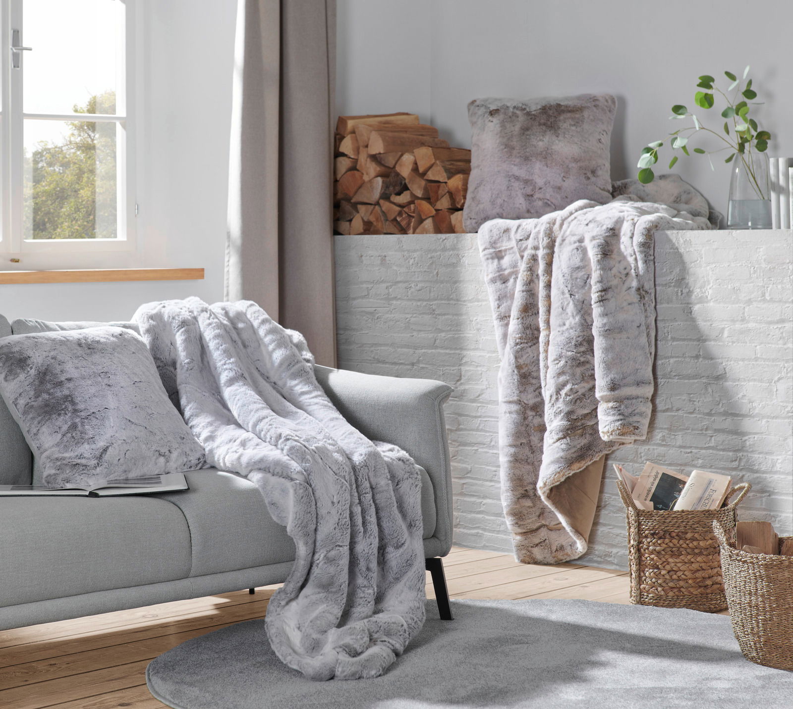 Gemütliche Wohnzimmer-Szene mit einer silberfuchsfarbenen Felldecke auf einem grauen Sofa, im Hintergrund ein Kamin mit Holzscheiten und Pflanzen, aus der Perspektive eines Betrachters im Raum.