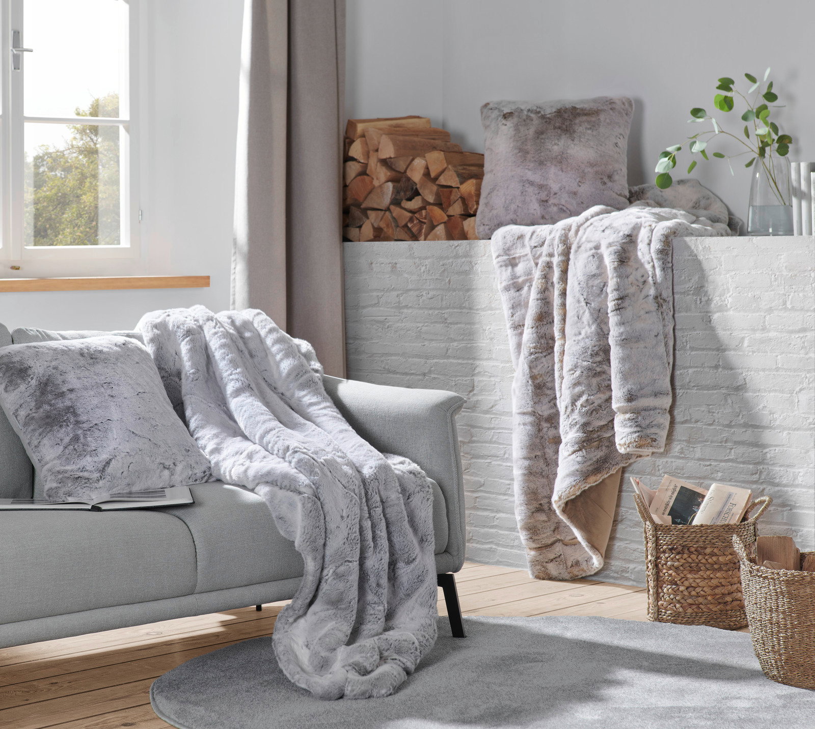 Gemütliche Wohnzimmer-Szene mit einer silberfuchsfarbenen Felldecke auf einem grauen Sofa, im Hintergrund ein Kamin mit Holzscheiten und Pflanzen, aus der Perspektive eines Betrachters im Raum.