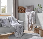 Gemütliche Wohnzimmer-Szene mit einer silberfuchsfarbenen Felldecke auf einem grauen Sofa, im Hintergrund ein Kamin mit Holzscheiten und Pflanzen, aus der Perspektive eines Betrachters im Raum.