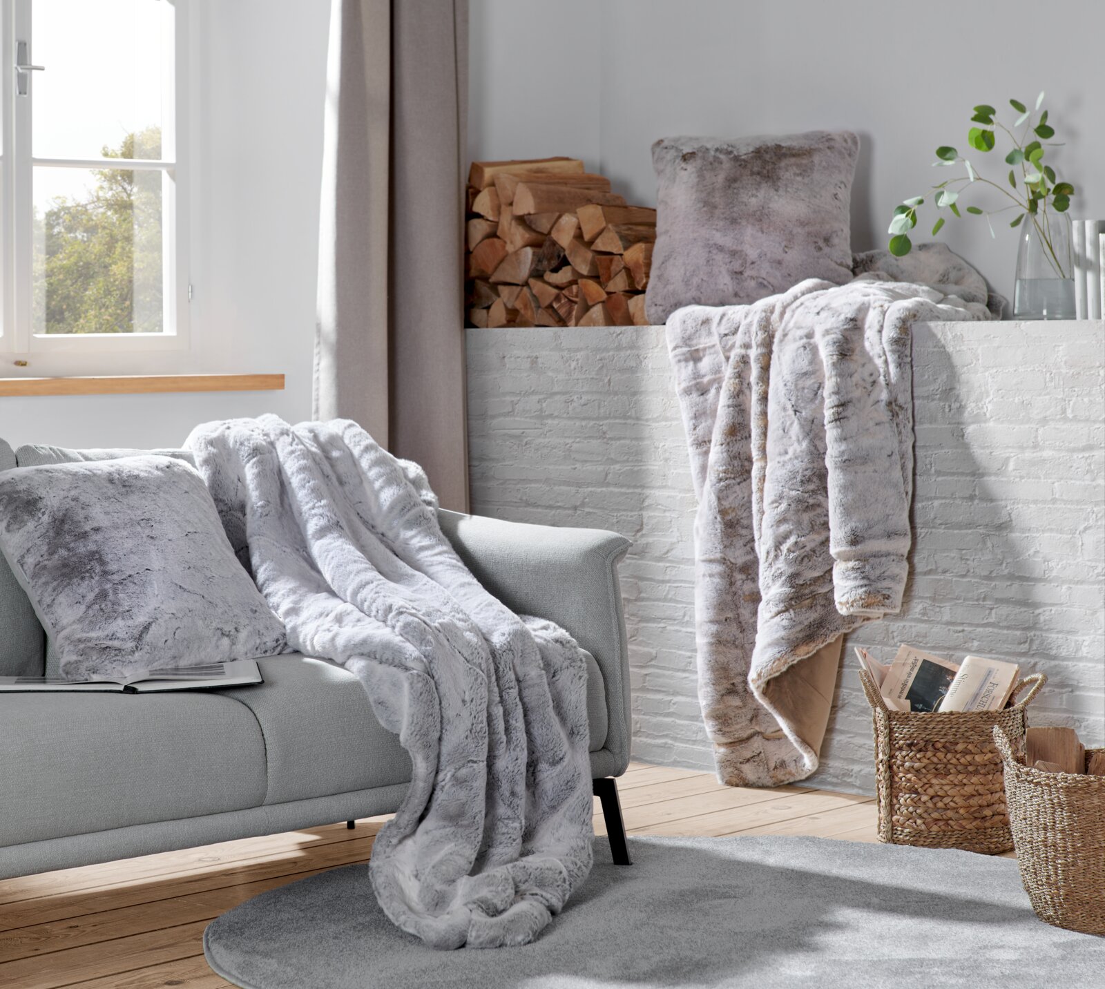 Gemütliche Wohnzimmer-Szene mit einer silberfuchsfarbenen Felldecke auf einem grauen Sofa, im Hintergrund ein Kamin mit Holzscheiten und Pflanzen, aus der Perspektive eines Betrachters im Raum.