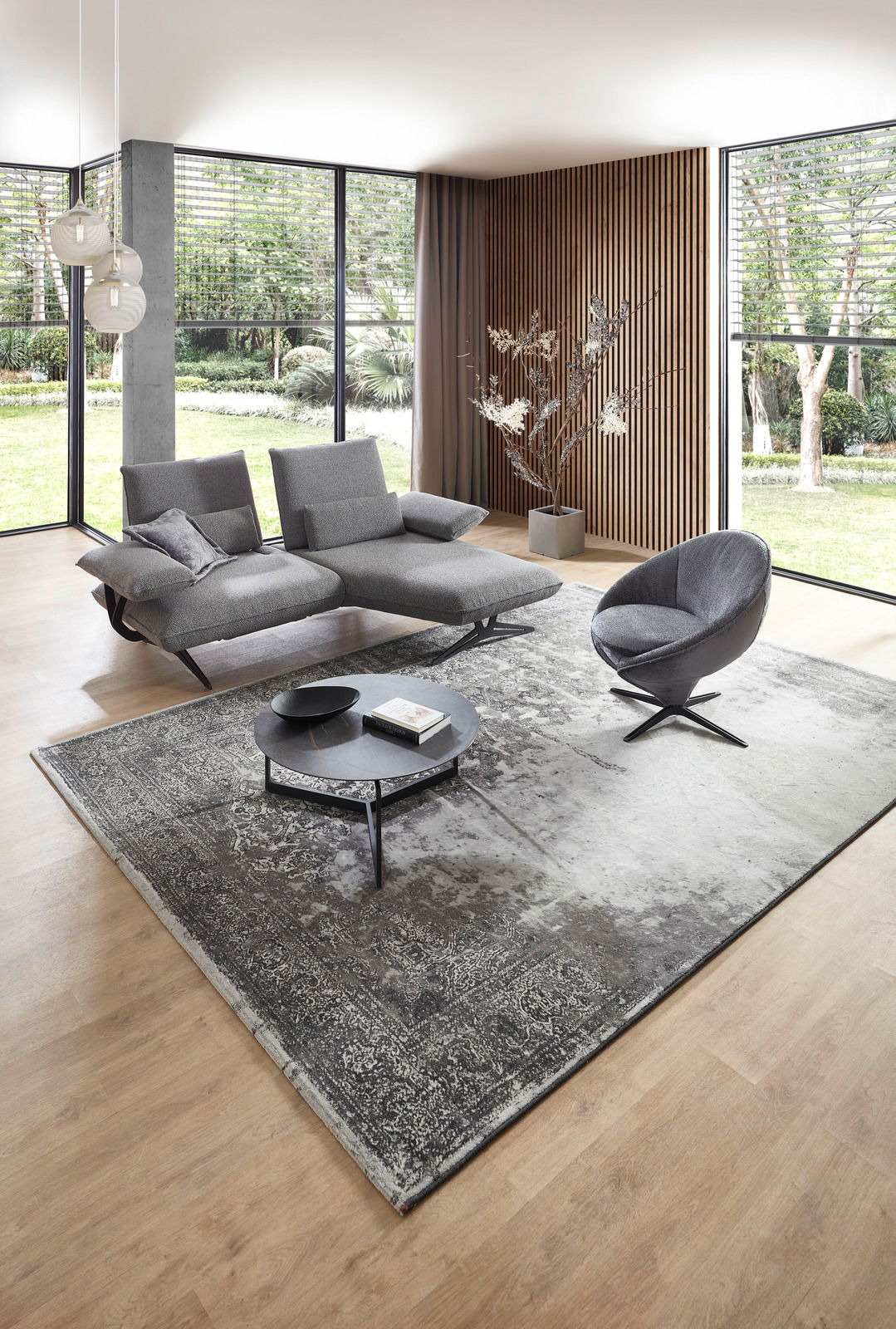 Moderner grauer Sessel in einem stilvollen Wohnzimmer mit großen Fenstern und Holzwandverkleidung, seitliche Perspektive.