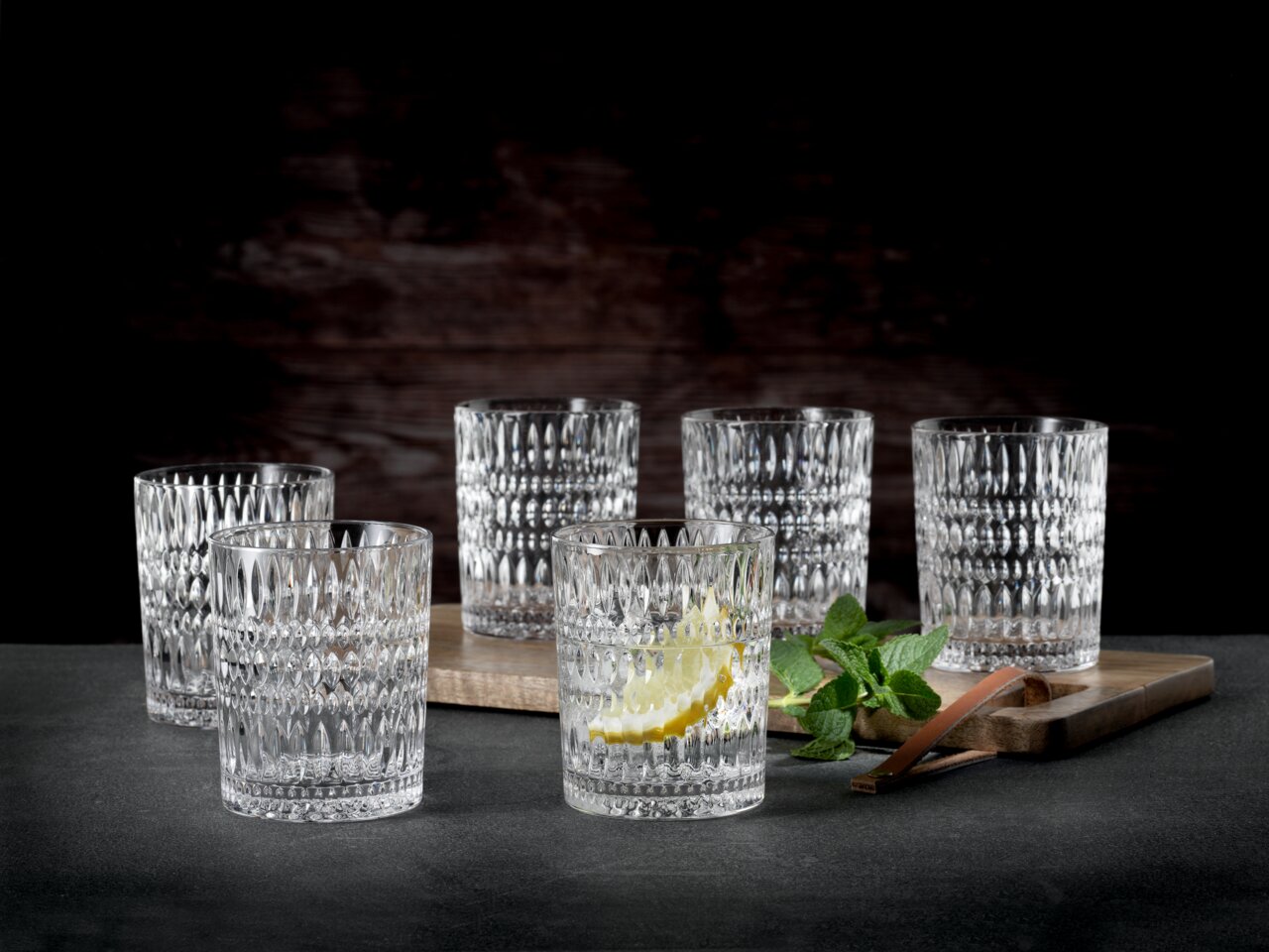 Nachtmann Becher ETHNO Sechs kristallklare Trinkgläser mit facettiertem Design, auf einem dunklen Hintergrund arrangiert, eines mit Zitronenscheibe und Minze, seitliche Perspektive.