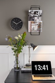 Wandmontierte Magazin-Aufbewahrung aus schwarzem Metall, frontal fotografiert, neben einer Uhr und über einem Schreibtisch mit Laptop und Blumen.