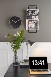 Wandmontierte Magazin-Aufbewahrung aus schwarzem Metall, frontal fotografiert, neben einer Uhr und über einem Schreibtisch mit Laptop und Blumen.