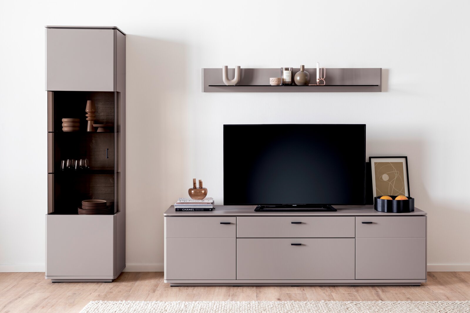 Modernes Wohnzimmer-Entertainment-Set mit grauem Lowboard, schwarzem Flachbild-TV, schwebendem Wandregal und hoher Vitrine mit Glasfront; Frontansicht auf hellem Holzboden.