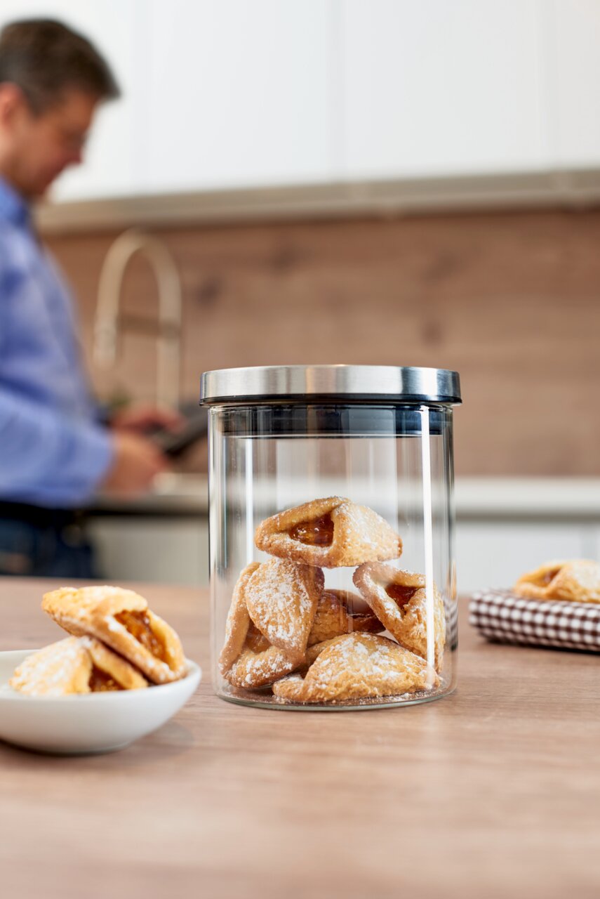 Glasdose Comodo 500ml mit Gebäck gefüllt, auf einem Holztisch in einer Küche platziert, im Hintergrund eine Person unscharf zu sehen.