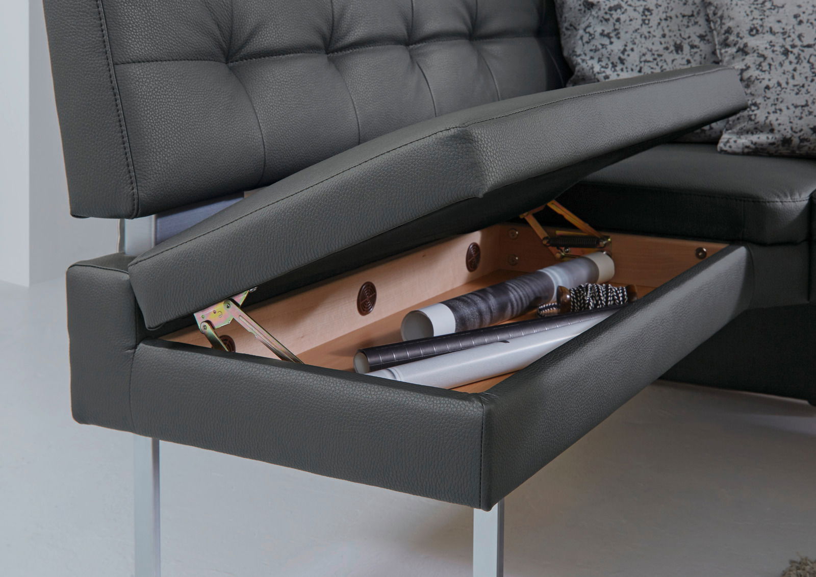 Nahaufnahme einer Eckbank mit aufklappbarem Stauraum unter der Sitzfläche, in dem verschiedene Gegenstände aufbewahrt werden. Perspektive von der Seite.