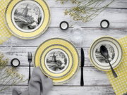 Draufsicht auf ein gedecktes Tisch-Arrangement mit gelb-weißem Geschirrset: Teller und Schalen mit schwarz-weißer Windmühlen-Landschaft, schwarzem Besteck, Gläsern und Teelichtern auf weißer Holzoberfläche.