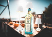 Akku-Tischleuchte VANNIE 3 in Weiß, präsentiert in einer sommerlichen Outdoor-Umgebung mit einer Flasche im Hintergrund. Perspektive: seitlich.