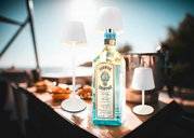 Akku-Tischleuchte VANNIE 3 in Weiß, präsentiert in einer sommerlichen Outdoor-Umgebung mit einer Flasche im Hintergrund. Perspektive: seitlich.