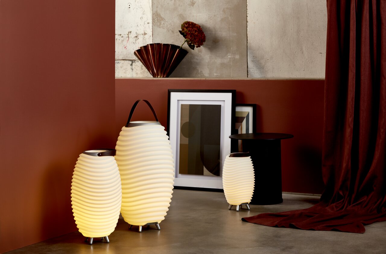 Drie golvende, verlichtende acculampen in verschillende maten, geplaatst op een betonnen vloer voor een roodbruine muur. De lampen hebben een handvat en staan in een gezellige woonomgeving. Perspectief: zijaanzicht.
