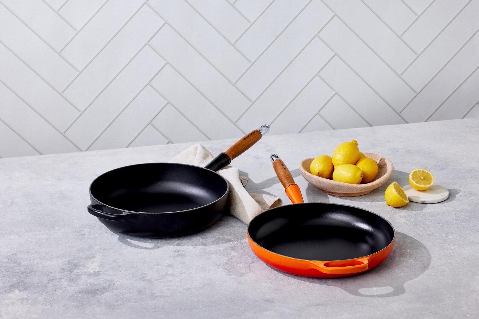 Zwei Bratpfannen auf einer Küchenarbeitsplatte, eine in Schwarz und eine in Ofenrot, mit Holzgriffen, aus der Vogelperspektive fotografiert. Daneben eine Schale mit Zitronen.