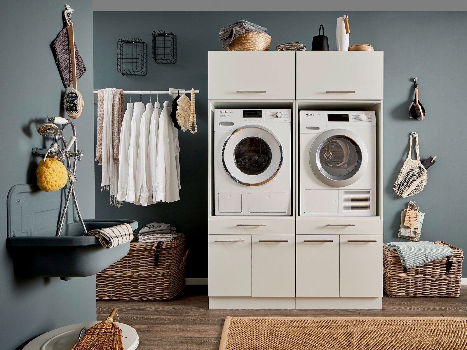Frontale Ansicht eines modernen Hauswirtschaftsraums mit zwei weißen Waschmaschinen und Trocknern in einem Schrank, umgeben von Körben und Hängeregalen.