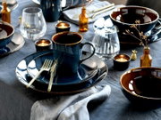Schwarzblaues Keramik-Geschirrset mit Tasse auf Untertasse und Teller sowie passenden Schalen, Gläsern, goldfarbenem Besteck und Teelichtern auf gedecktem Tisch, aus leicht erhöhter seitlicher Perspektive