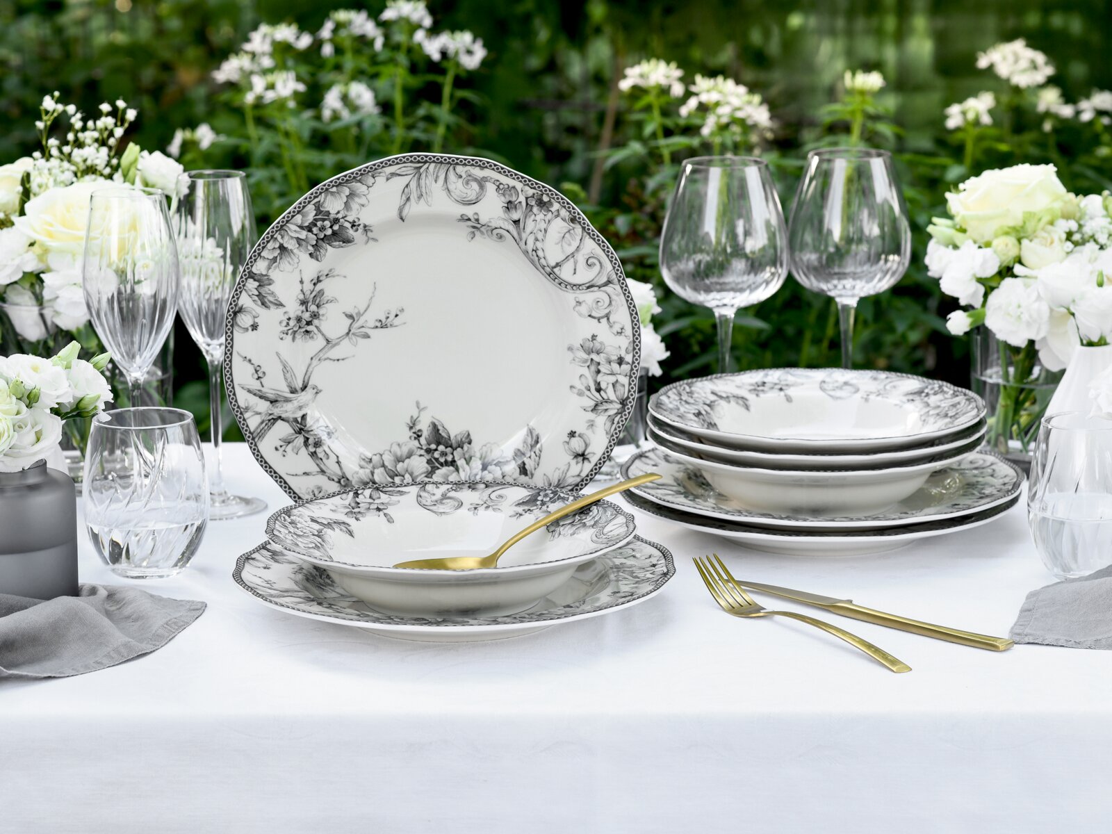 Elegantes weißes Porzellan-Geschirrset mit schwarzem floralen Dekor (Teller und Schalen) auf gedecktem Tisch im Garten, frontal aus leichter Schrägansicht fotografiert.