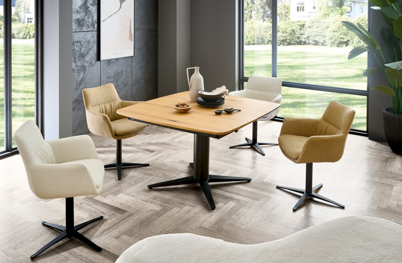 Moderner Essbereich mit vier Armlehnstühlen um einen quadratischen Holztisch, umgeben von großen Fenstern mit Blick ins Grüne. Perspektive: seitlich.