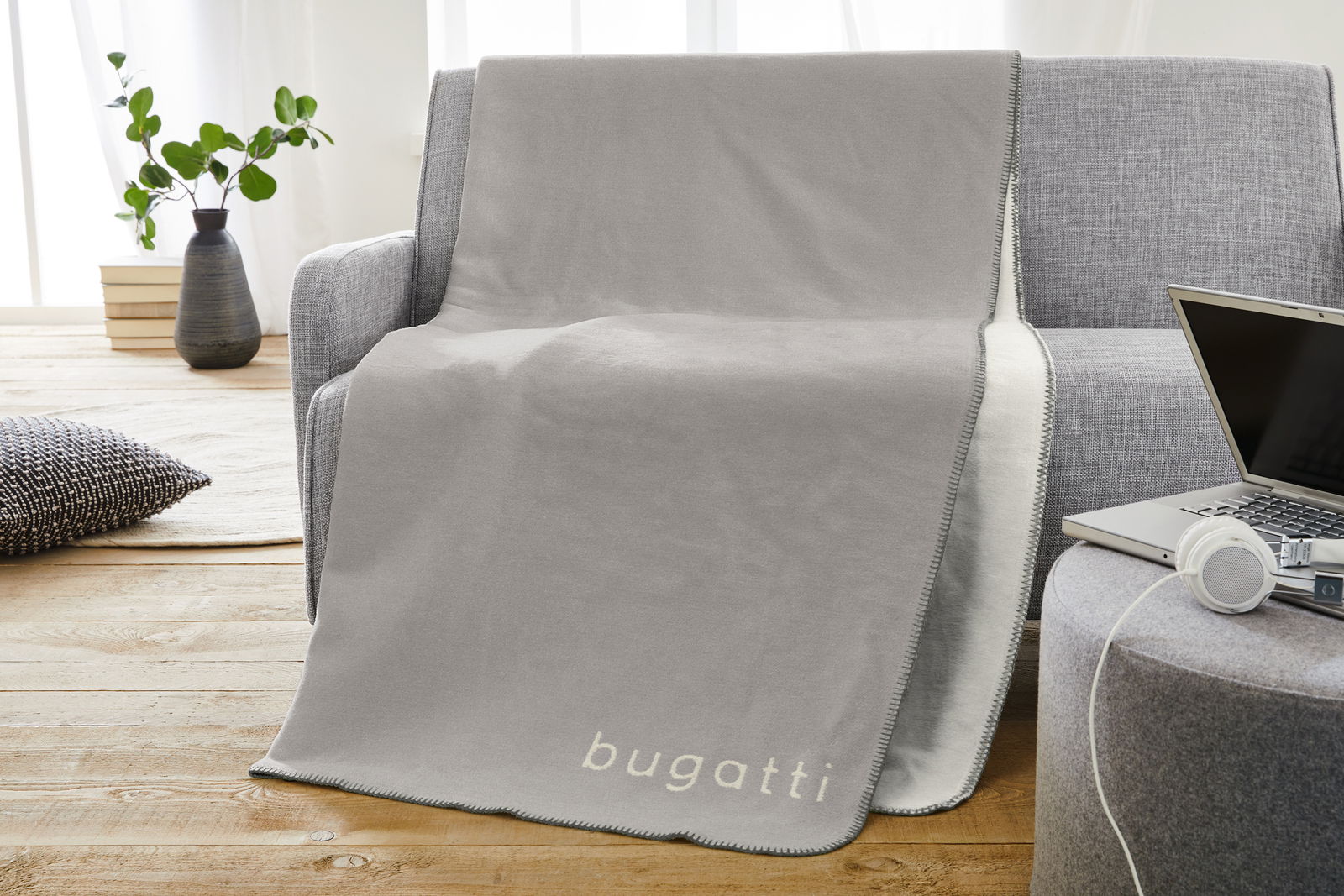 Graue Jacquard Decke 150x200cm von bugatti, drapiert über einem grauen Sofa in einem modernen Wohnzimmer mit Holzboden und Pflanzen im Hintergrund, seitliche Perspektive.