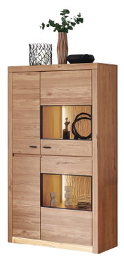 Frontale Ansicht einer hohen, modernen Vitrine/Kommode aus hellem Massivholz mit zwei beleuchteten offenen Fächern und schwarzen Griffen; oben stehen Dekoartikel und eine Vase mit Grünpflanze.