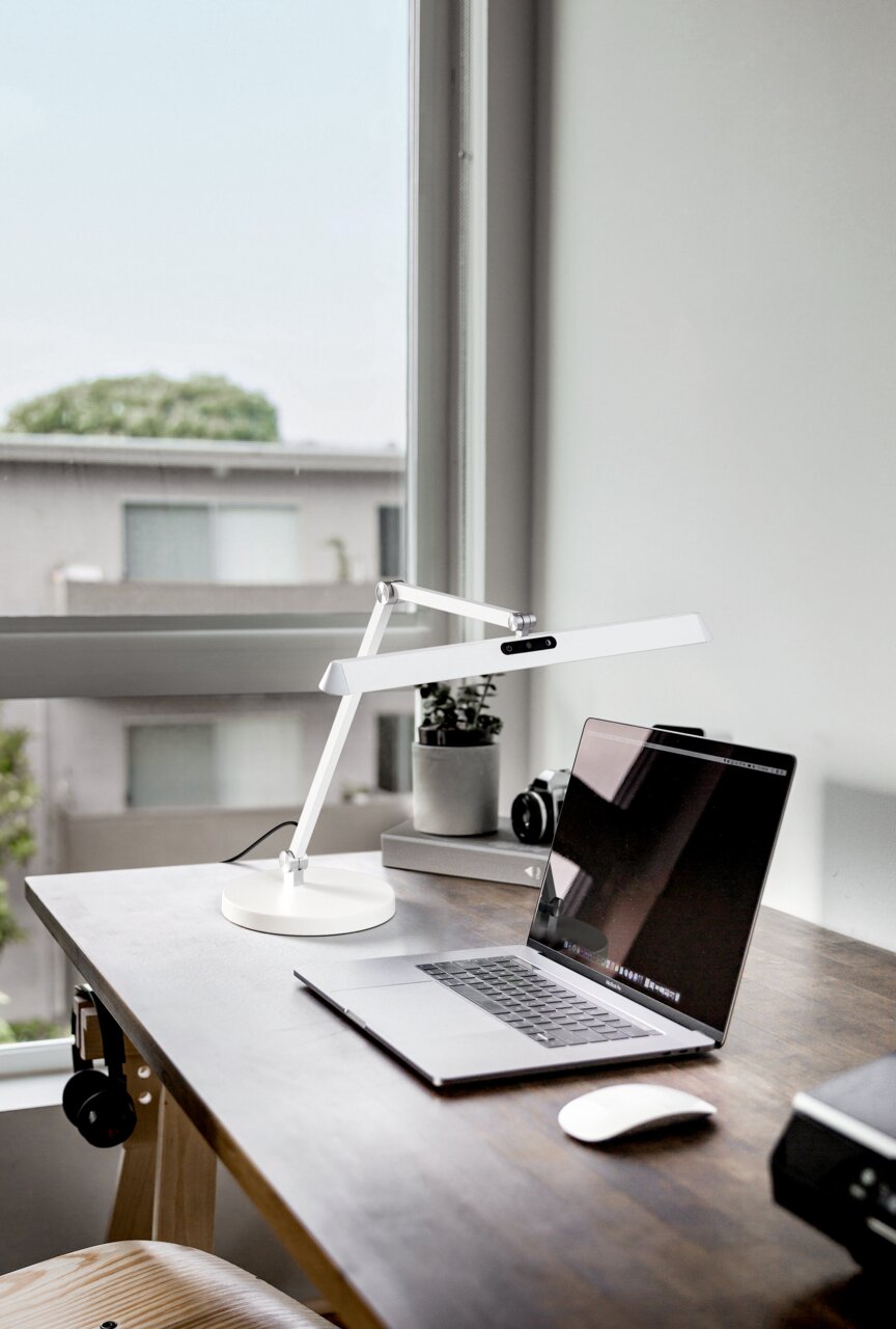 Moderne witte tafellamp Beba op een bureau naast een laptop, zijaanzicht.