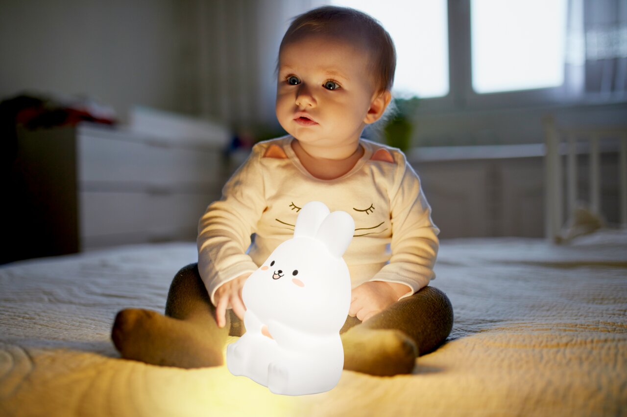 Ein Baby sitzt auf einem Bett und hält ein leuchtendes Nachtlicht in Form eines Hasen in den Händen. Das Zimmer ist sanft beleuchtet, und das Nachtlicht strahlt ein warmes Licht aus. Perspektive von vorne.