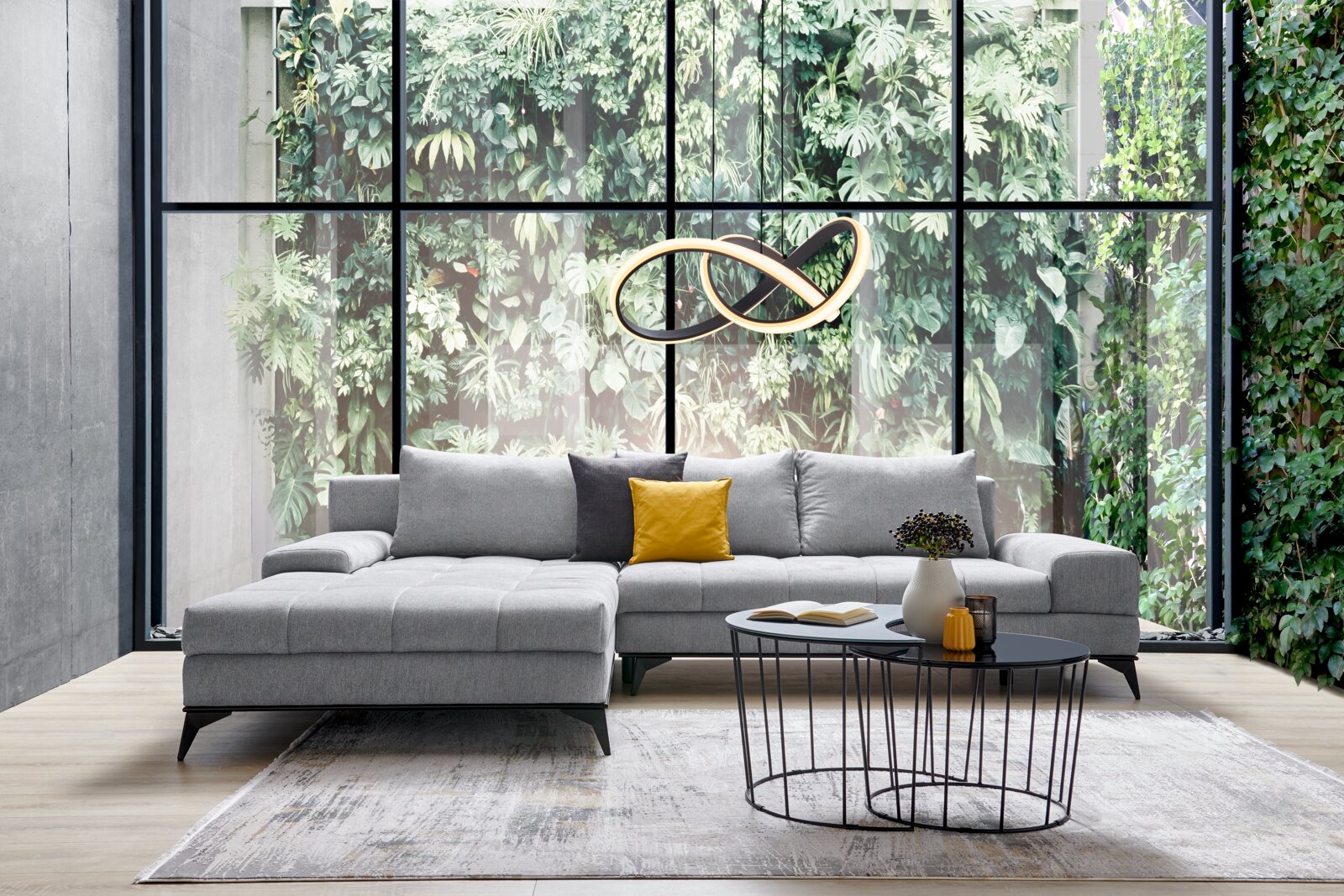 Z2 Ecksofa FIGARO Graue Rundecke Ottli in einem modernen Wohnzimmer mit großen Fenstern und grüner Pflanzenwand, frontal betrachtet.