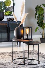 Zwei runde Beistelltische aus Metall mit schwarzer Oberfläche, dekoriert mit einer schwarzen Vase und einer modernen Lampe, seitliche Perspektive.