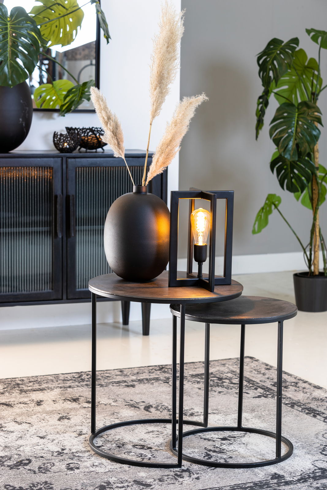 Zwei runde Beistelltische aus Metall mit schwarzer Oberfläche, dekoriert mit einer schwarzen Vase und einer modernen Lampe, seitliche Perspektive.