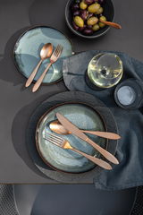Vogelperspektive auf ein stilvoll gedecktes Tischarrangement mit modernem Besteck in Kupferoptik auf blauen Tellern, begleitet von einem Glas Weißwein und einer Schale mit Oliven.
