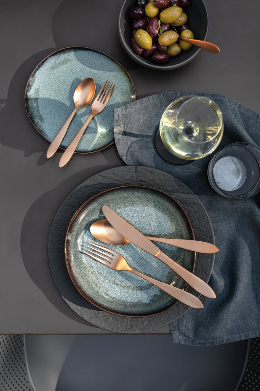 Villeroy & Boch Tafelbesteck 20-tlg. MANUFACTURE ROCK CUTLERY Vogelperspektive auf ein stilvoll gedecktes Tischarrangement mit modernem Besteck in Kupferoptik auf blauen Tellern, begleitet von einem Glas Weißwein und einer Schale mit Oliven.