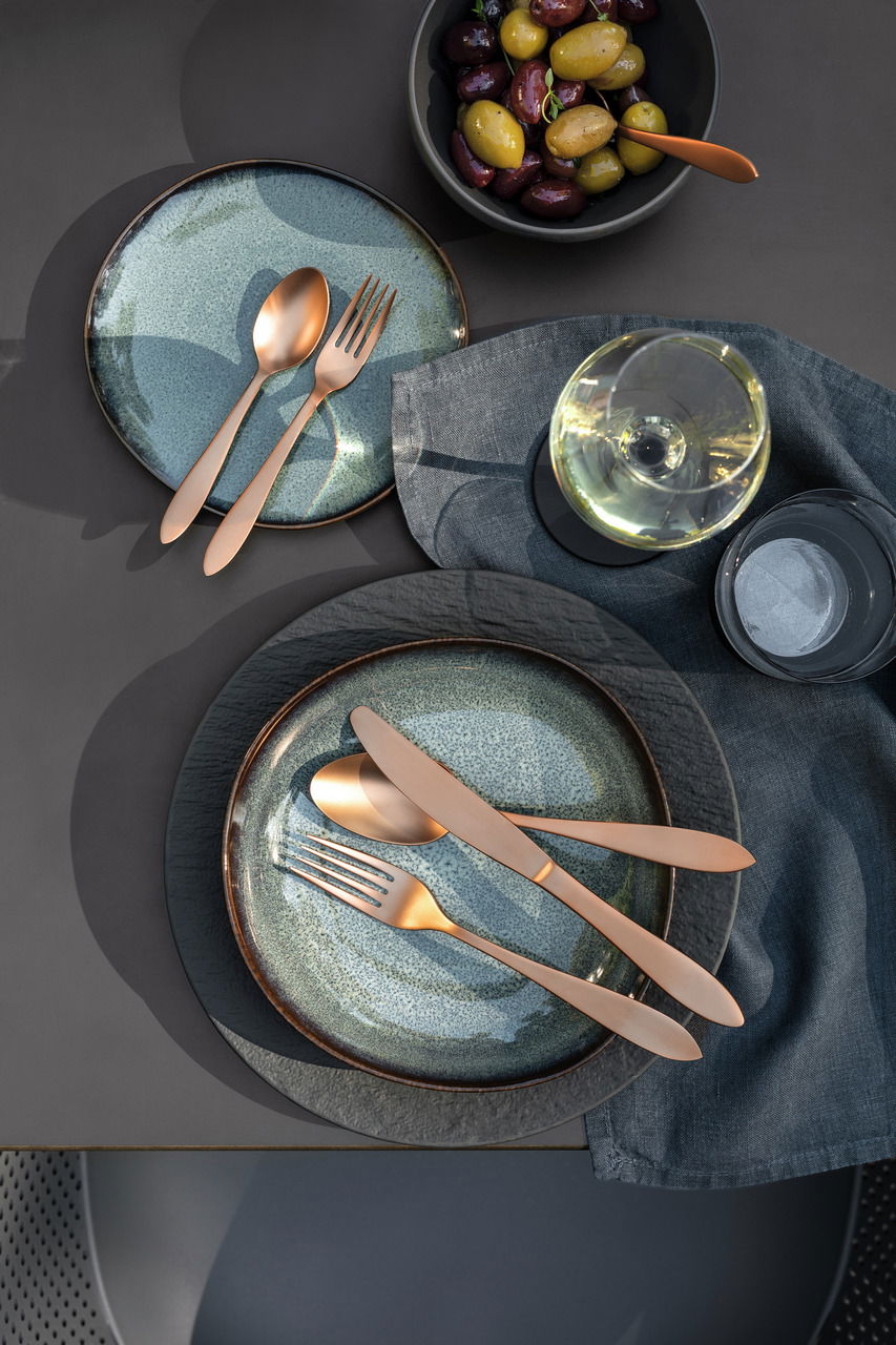 Vogelperspektive auf ein stilvoll gedecktes Tischarrangement mit modernem Besteck in Kupferoptik auf blauen Tellern, begleitet von einem Glas Weißwein und einer Schale mit Oliven.