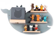 Graues, wal-/fischförmiges Wandregal aus Holz mit drei kleinen Ablageleisten und einer größeren Leiste, darauf bunte Spielfiguren und ein grauer Kinderlautsprecher mit Katzenmotiv; Frontansicht.