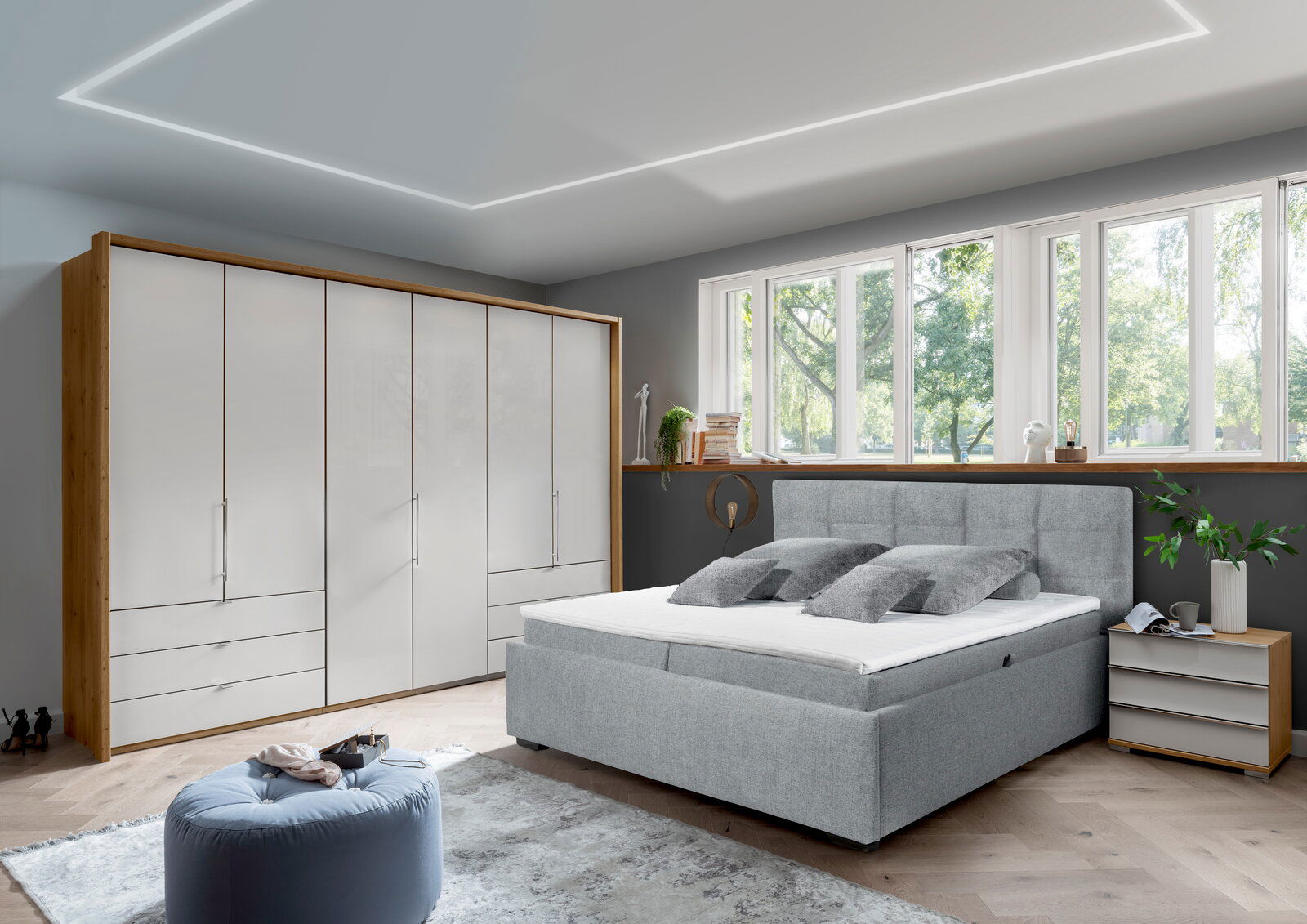 Modernes Schlafzimmer mit einem grauen Polsterbett in Frontalansicht, umgeben von einem großen Kleiderschrank, einem Nachttisch und einem großen Fenster mit Blick ins Grüne.