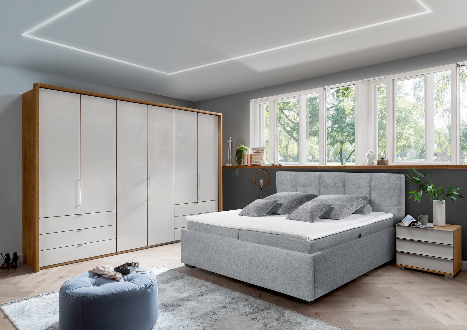 Modernes Schlafzimmer mit einem grauen Polsterbett in Frontalansicht, umgeben von einem großen Kleiderschrank, einem Nachttisch und einem großen Fenster mit Blick ins Grüne.