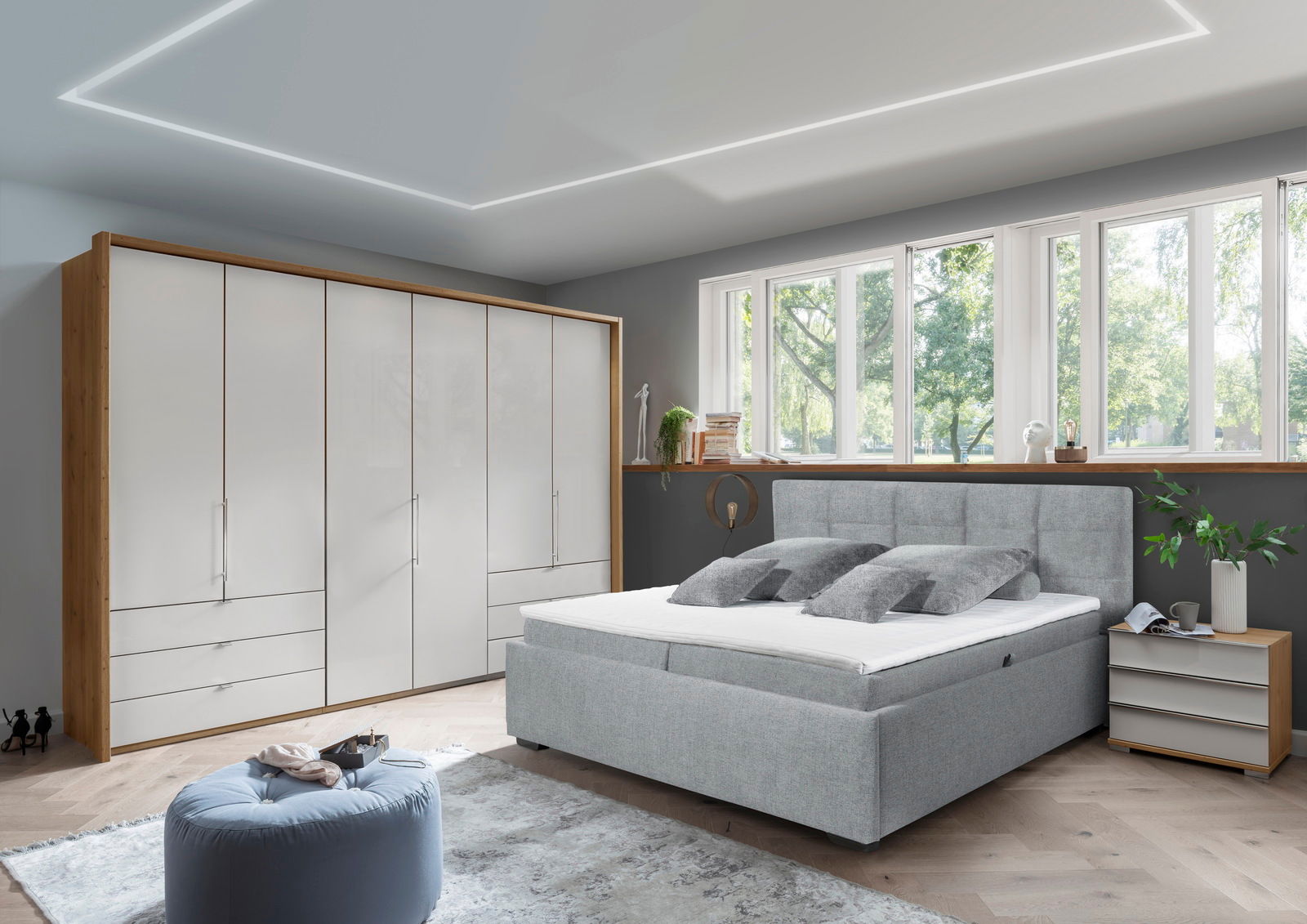 Modernes Schlafzimmer mit einem grauen Polsterbett in Frontalansicht, umgeben von einem großen Kleiderschrank, einem Nachttisch und einem großen Fenster mit Blick ins Grüne.